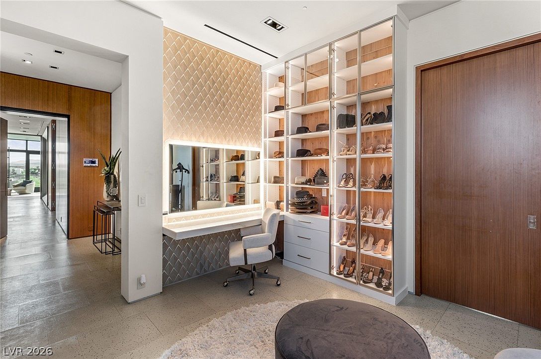 This luxurious walk-in closet features a dedicated vanity area with a large, illuminated mirror and a modern desk chair. To the right, a custom shelving unit with glass-front doors provides organized storage for shoes and accessories, accented by integrated lighting. The space is finished with a neutral color palette, patterned wall detailing, and a plush circular ottoman in the foreground, creating a sophisticated and functional dressing area.