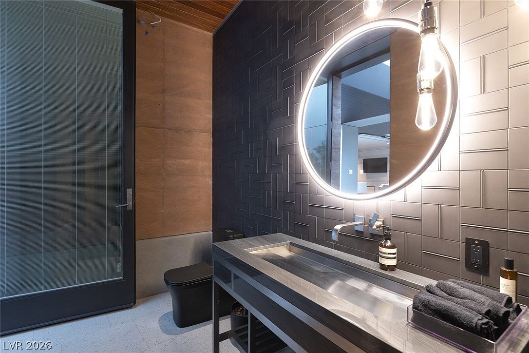 This modern powder room features a striking dark aesthetic with textured, geometric wall tiles and a sleek, trough-style sink set on a minimalist metal frame. A large, circular backlit mirror serves as the focal point, complemented by industrial-style pendant lighting and a dark-toned toilet. The space is framed by a tall glass door and warm wood-toned ceiling accents, creating a sophisticated and moody atmosphere.