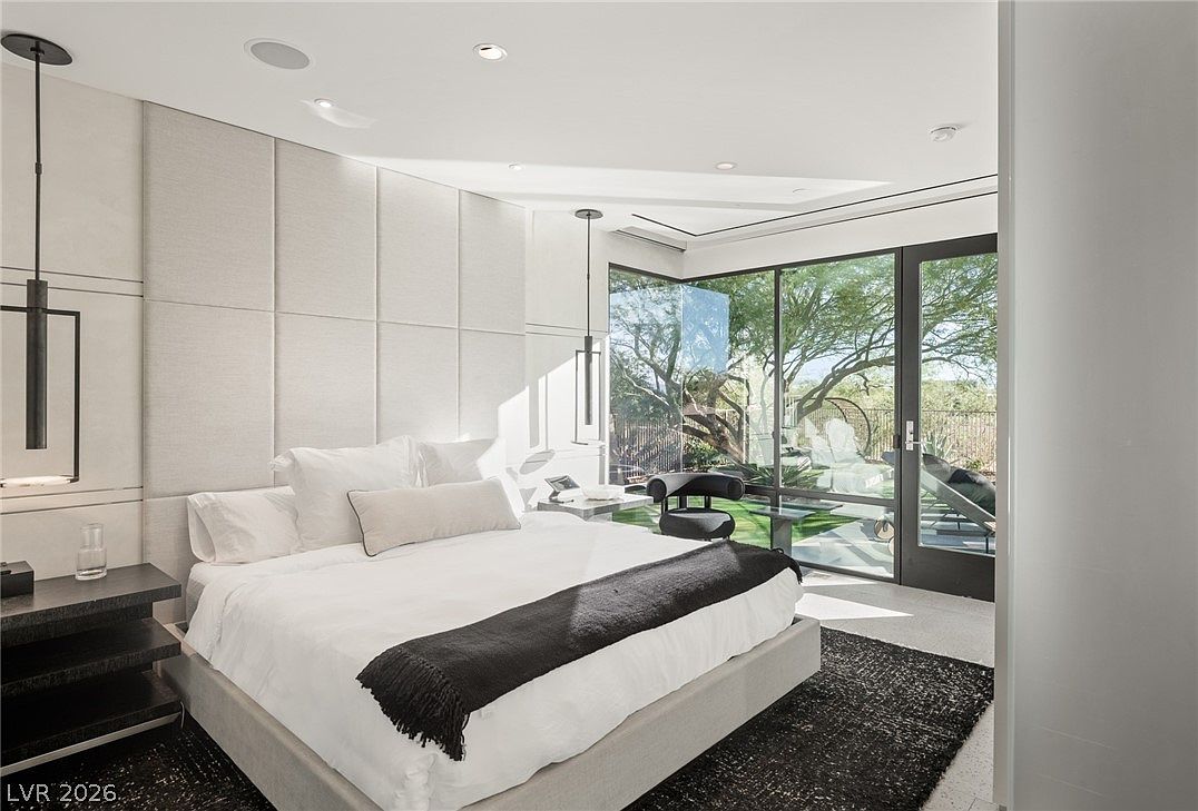 This modern primary bedroom features a large bed with a plush white duvet and a dark throw, set against a wall with large, upholstered rectangular panels. The room is bathed in natural light from expansive floor-to-ceiling windows that offer a clear view of the outdoor landscaping. A dark, textured area rug anchors the space, while minimalist pendant lights hang on either side of the bed, contributing to the sleek, contemporary aesthetic.