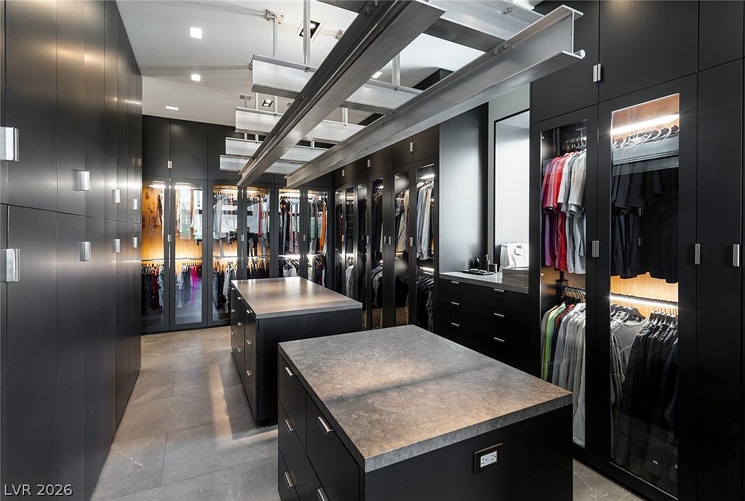 This expansive, modern walk-in closet features dark cabinetry with glass-front doors and integrated lighting, creating a sophisticated and organized storage space. Two central islands with stone-look countertops provide additional surface area, while a unique industrial-style metal beam structure is suspended from the ceiling. The room is finished with large-format floor tiles, offering a sleek and high-end aesthetic.