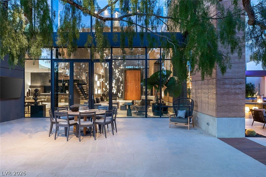 This sophisticated outdoor patio space features a dining set arranged on light-toned stone pavers, set against a backdrop of expansive floor-to-ceiling glass walls that reveal a luxurious interior. The area is shaded by the overhanging branches of a mature tree, creating a serene atmosphere that blends indoor and outdoor living. A modern lounge chair sits to the side, while the overall architectural style emphasizes clean lines, transparency, and high-end contemporary design.