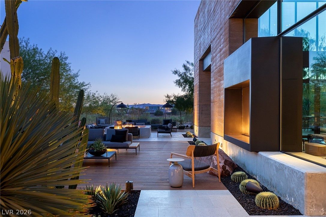 This modern outdoor living space features a spacious wooden deck furnished with contemporary lounge seating and a central fire pit. A striking architectural element includes a dark, cantilevered fireplace structure attached to the side of the home, complemented by desert landscaping with cacti and stone accents. The perspective captures a serene twilight atmosphere, highlighting the warm glow of the lighting against the clean lines of the building.