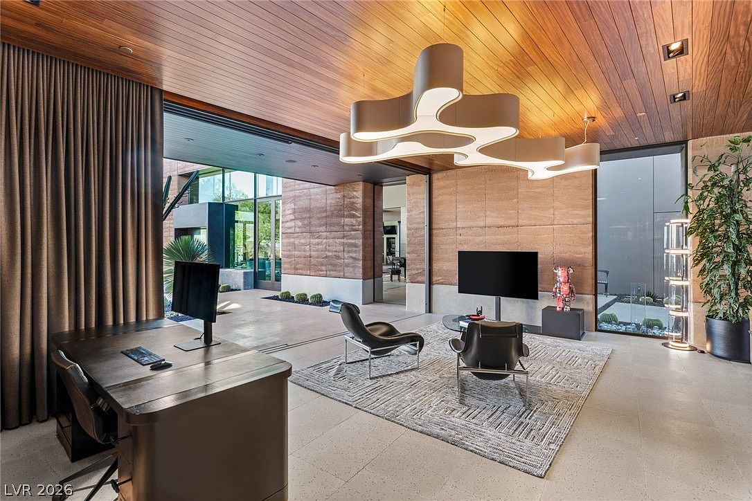 This sophisticated home office features a seamless transition to an outdoor patio through expansive sliding glass walls, creating an indoor-outdoor living experience. The room is anchored by a dark wood desk and a seating area with two leather chairs atop a textured area rug, all illuminated by a unique, sculptural modern chandelier. The space is defined by warm wood-paneled ceilings and a textured stone wall, offering a luxurious and contemporary aesthetic.