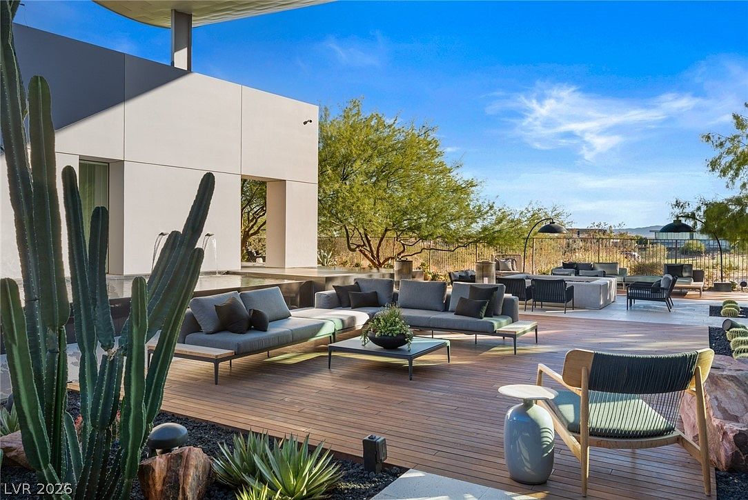 This expansive outdoor living space features a modern wooden deck furnished with comfortable gray sectional seating and a central coffee table. A sleek, minimalist white structure with a water feature provides a sophisticated backdrop, while desert landscaping and tall cacti frame the scene. The area is designed for luxury entertaining, offering a seamless transition between indoor and outdoor living under a clear blue sky.