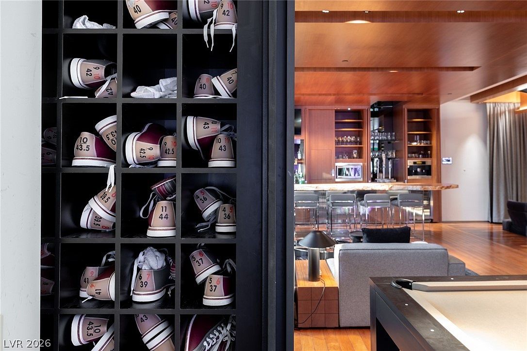 This image features a custom-built, dark-toned shelving unit designed for shoe storage, prominently displayed in the foreground. Through the open doorway, a sophisticated living area is visible, complete with a bar, bar stools, and a pool table, suggesting a high-end entertainment space. The perspective creates a transition from a functional storage area into a luxurious, open-concept interior.