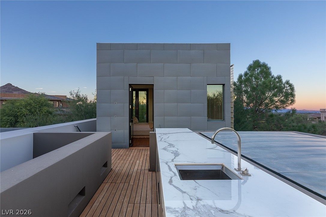 This rooftop deck features a sleek outdoor kitchen area with a stone-topped counter and a modern faucet. A wooden plank deck leads to a doorway opening into a minimalist structure clad in large, rectangular panels. The setting offers an elevated perspective with views of the surrounding landscape and distant mountains under a clear sky.