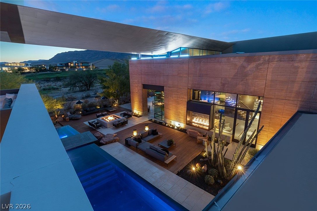 This high-angle perspective captures a luxurious modern outdoor living space at dusk, featuring a multi-level deck with a swimming pool and a fire pit lounge area. The architecture showcases clean lines, warm exterior lighting, and large glass walls that connect the indoor and outdoor environments. A desert-inspired landscape with cacti and stone accents complements the contemporary design, creating a sophisticated and serene atmosphere.