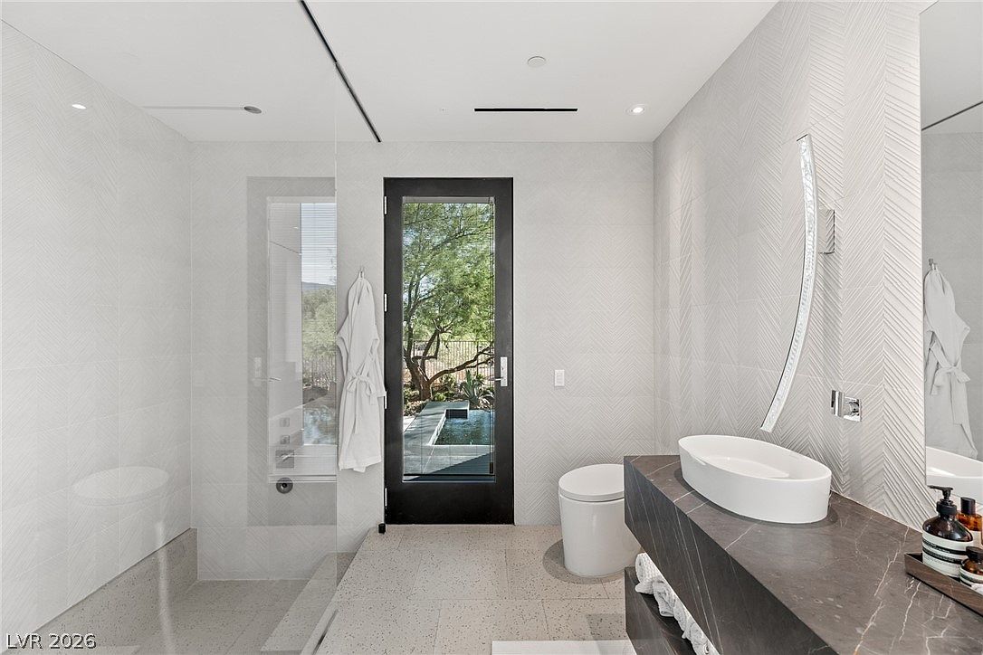 This modern primary bathroom features a sleek, dark stone vanity with a vessel sink and a large, curved wall-mounted mirror. To the left, a glass-enclosed shower area is visible, while a minimalist toilet sits near a black-framed glass door that opens to an outdoor view. The room is finished with light-toned, textured wall tiles and neutral floor tiles, creating a clean and sophisticated atmosphere.