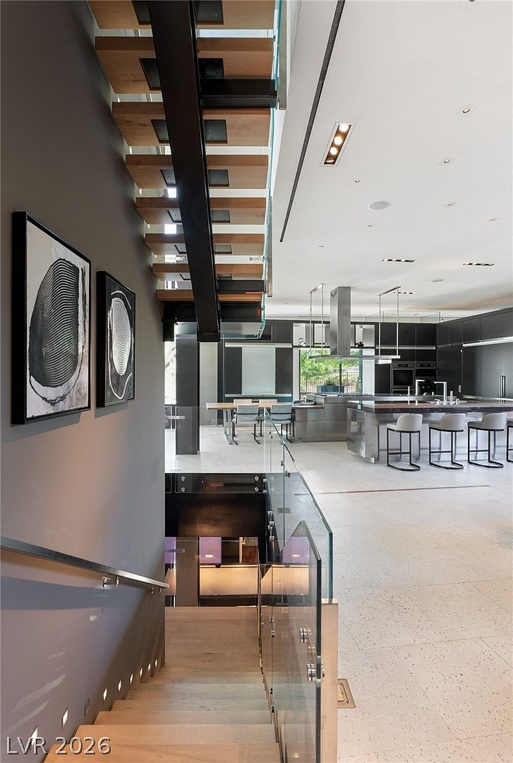 This perspective captures a dramatic, modern staircase with light wood treads and a glass railing, descending into an open-concept living area. The view highlights the architectural contrast between the dark, moody wall adorned with abstract art and the bright, expansive kitchen space in the background. The clean lines and minimalist aesthetic create a sophisticated, high-end atmosphere.