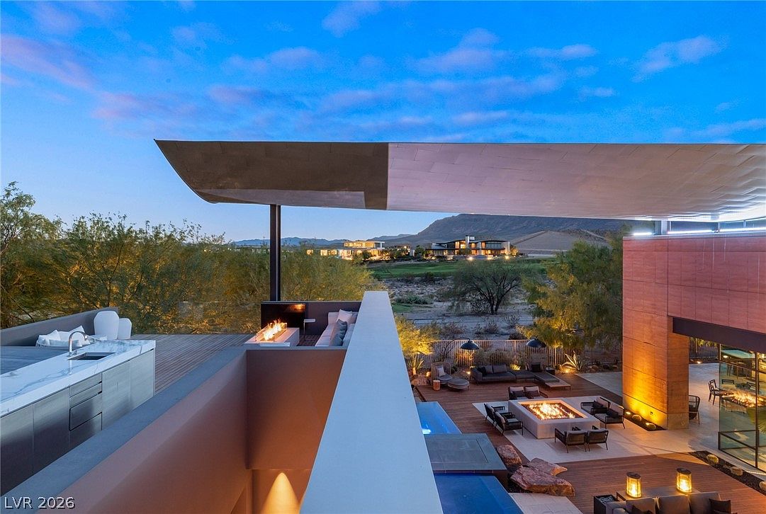 This high-angle shot captures a luxurious multi-level outdoor living space at dusk, featuring a sleek, modern architectural design with a prominent cantilevered roofline. The scene includes an upper-level terrace with a linear fire feature and a lower-level deck equipped with a fire pit, lounge seating, and a swimming pool. The expansive view overlooks a desert landscape with distant mountains, creating a sophisticated and serene atmosphere.