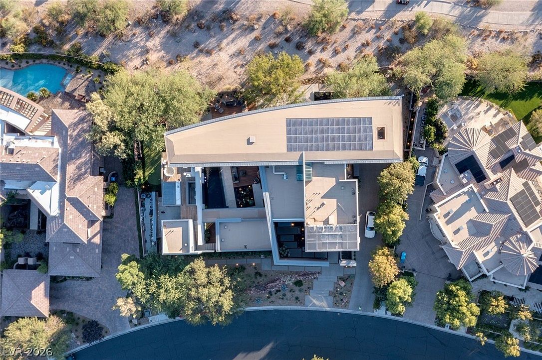 This high-angle aerial perspective captures a modern, flat-roofed residence situated within a desert-landscaped neighborhood. The home features a prominent solar panel array on the roof, a private swimming pool, and a multi-level outdoor living area with integrated patio spaces. The surrounding property includes manicured lawns, mature trees, and a curved driveway, showcasing a contemporary architectural design.