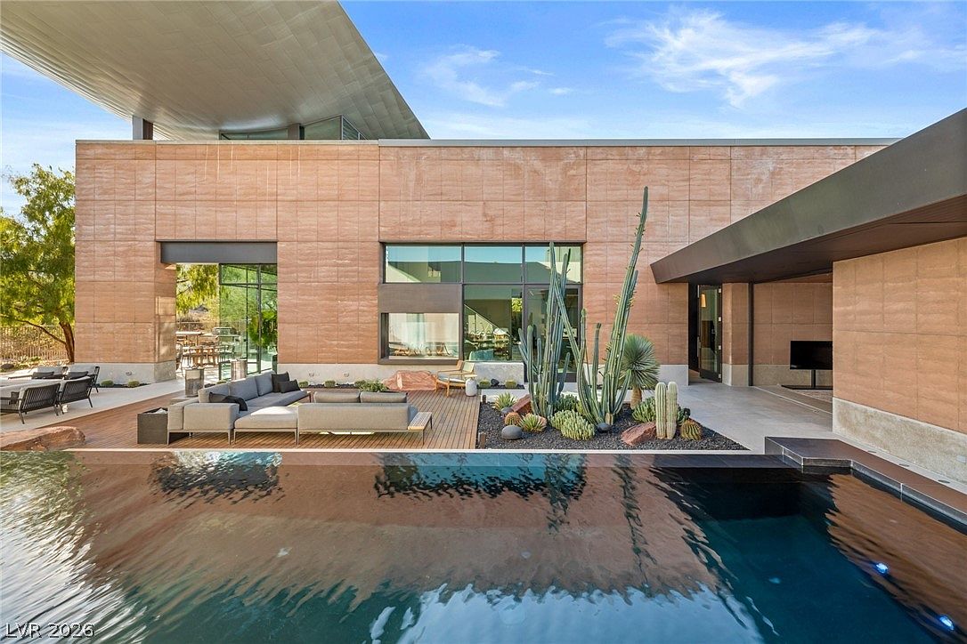 This modern desert estate features a sleek, dark-tiled infinity pool that reflects the clear blue sky and the architectural lines of the home. A spacious wooden deck provides an inviting outdoor lounge area, complemented by a landscaped garden bed filled with various cacti and desert flora. The home's exterior is defined by large, clean-lined stone panels and expansive glass walls that seamlessly connect the indoor and outdoor living spaces.