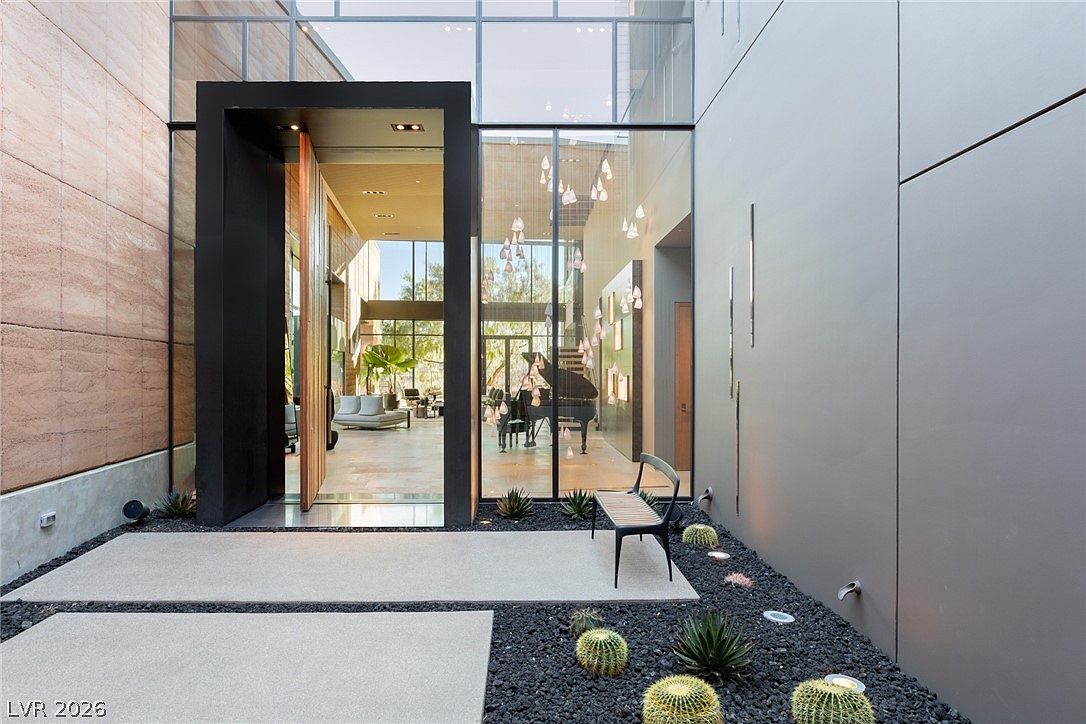 This striking modern entryway features a massive, oversized wooden pivot door framed by a bold black surround, set against a backdrop of expansive glass walls. The exterior courtyard is landscaped with dark gravel, desert cacti, and a minimalist bench, creating a sophisticated and architectural aesthetic. The perspective looks directly into the bright, open-concept interior, where a grand piano and contemporary furnishings are visible through the glass.