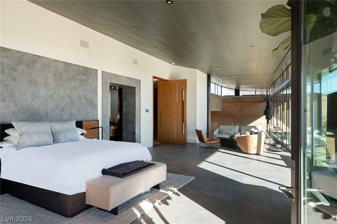 This expansive primary bedroom features a modern aesthetic with a large bed centered against a textured gray wall. The room transitions into a seating area with a lounge chair and sofa, all illuminated by natural light streaming through extensive glass walls that offer an indoor-outdoor feel. A unique, dark-toned ceiling adds architectural interest, while the open-concept layout flows seamlessly toward a private bathroom entrance.