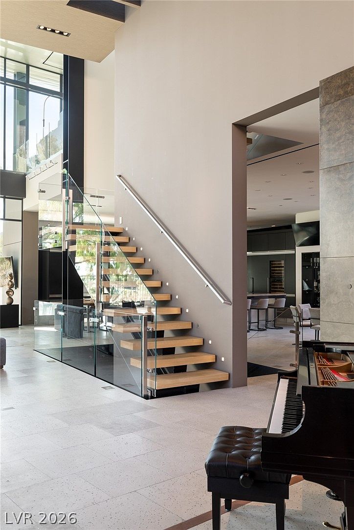 This modern, open-concept interior features a striking floating staircase with light wood treads and a glass railing system. The perspective looks from a tiled foyer toward a kitchen area in the background, with a grand piano positioned in the foreground. The space is defined by high ceilings, neutral wall tones, and large windows that allow for abundant natural light.