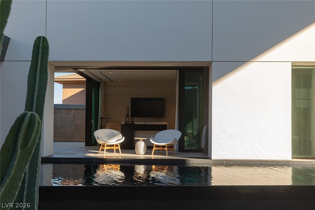 This image captures a modern indoor-outdoor living space featuring a sleek, dark-tiled swimming pool in the foreground. Beyond the pool, a covered patio area is furnished with two white lounge chairs and a small side table, leading into a minimalist living room visible through a wide opening. The architectural style is contemporary, characterized by clean lines, smooth light-colored walls, and a tall cactus framing the left side of the composition.