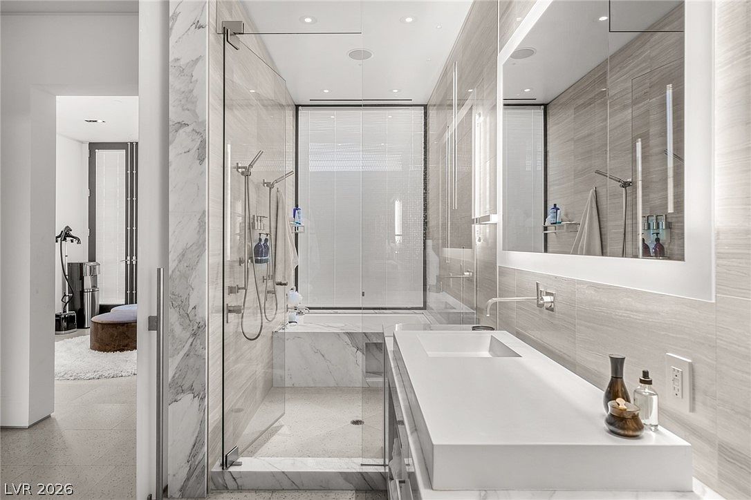 This modern primary bathroom features a sleek, wall-mounted vanity with a rectangular sink and a large, frameless mirror. A glass-enclosed wet room area contains both a shower and a bathtub, all finished with light-toned, veined stone surfaces. The perspective looks from the vanity area toward the wet room, highlighting the clean lines and contemporary design of the space.