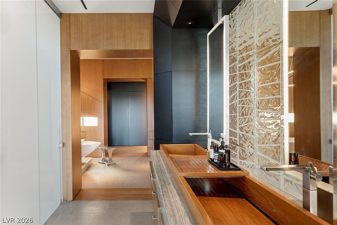 This modern primary bathroom features a unique wooden trough-style sink set into a stone vanity, complemented by a textured glass block wall with integrated lighting. The space transitions into a warm, wood-paneled hallway that leads to a secondary area containing a soaking tub. The design aesthetic is sophisticated and minimalist, utilizing a blend of natural wood tones and sleek, dark architectural elements.
