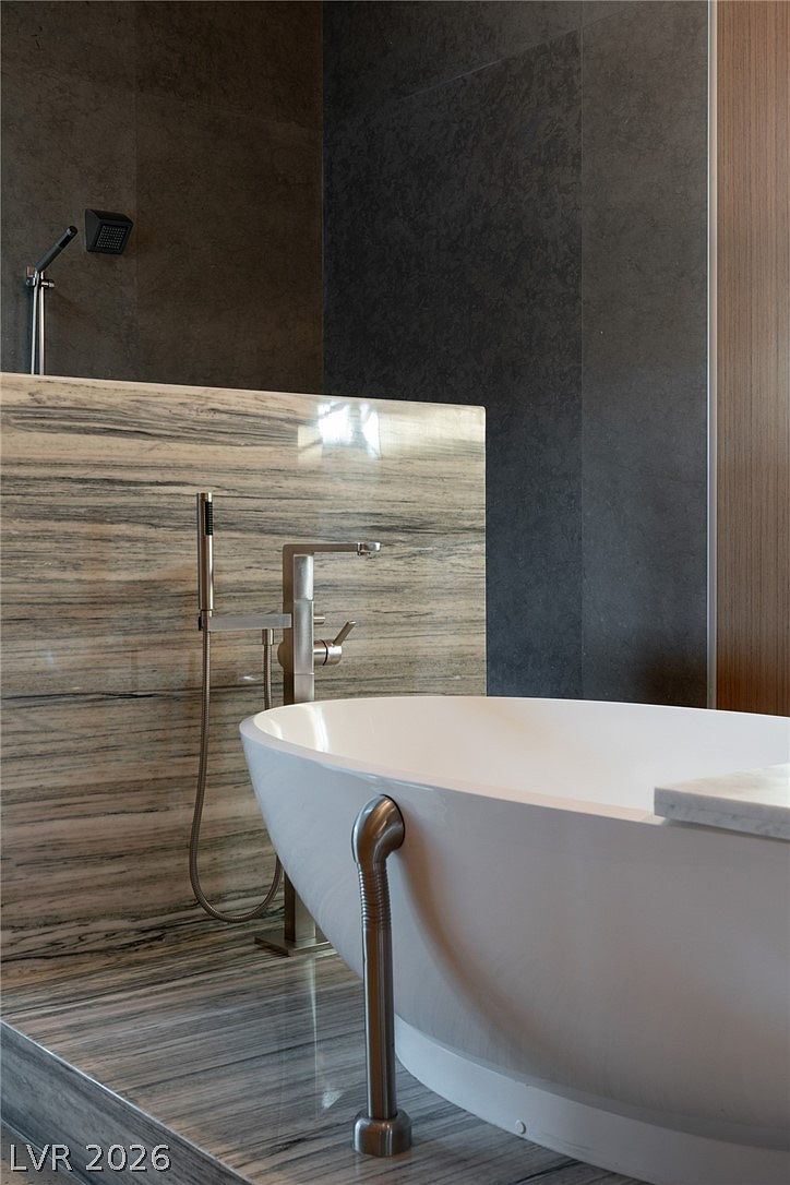 This close-up perspective highlights a luxurious freestanding soaking tub set against a backdrop of horizontally veined stone wall cladding. A sleek, modern brushed-metal faucet assembly stands beside the tub, complemented by a matching handheld shower fixture. The composition emphasizes the high-end materials and clean, contemporary design of the bathroom space.