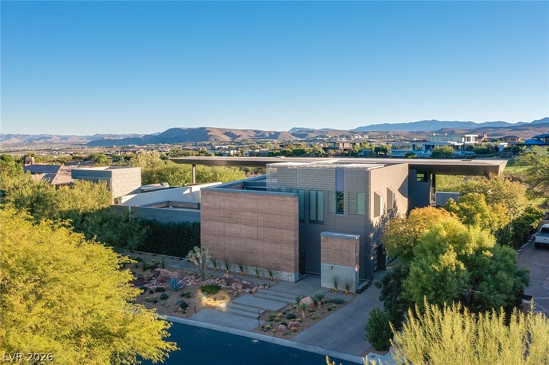 This aerial perspective captures a striking contemporary residence characterized by its clean lines, flat rooflines, and a mix of horizontal wood-toned siding and smooth concrete surfaces. The home is nestled in a desert landscape with native vegetation and a paved driveway leading to the entrance, all set against a backdrop of rolling mountains. The architectural design emphasizes a modern, minimalist aesthetic that integrates seamlessly with the surrounding arid environment.