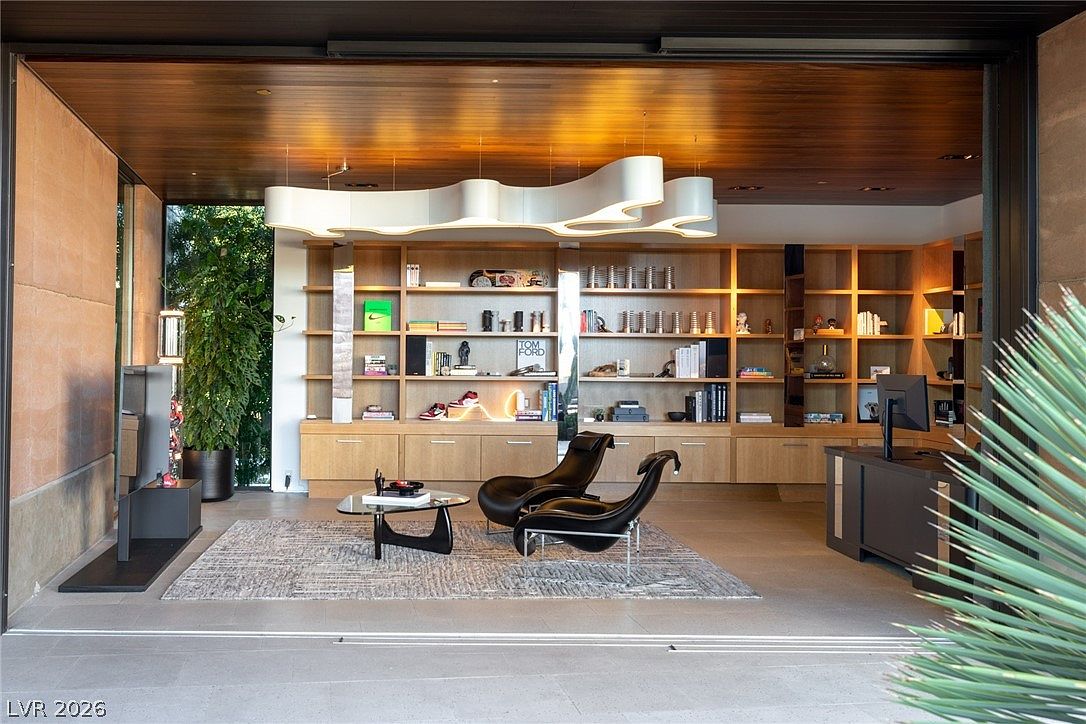 This sophisticated home office features a seamless transition to the outdoors through a wide, open wall, creating an airy and expansive atmosphere. The room is anchored by a custom light wood shelving unit that spans the back wall, displaying curated decor and books, while two sleek black lounge chairs sit atop a textured area rug. A unique, sculptural light fixture hangs from the warm wood-paneled ceiling, adding a modern artistic touch to the refined, professional space.