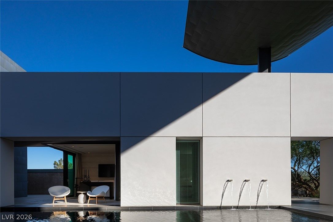 This modern architectural exterior features a sleek, minimalist design with smooth white wall panels and a striking cantilevered roof structure. A tranquil infinity-edge pool occupies the foreground, accented by three elegant water spouts that create a rhythmic, calming effect. The scene includes a partially shaded patio area furnished with contemporary white chairs, offering a seamless transition between indoor and outdoor living spaces.