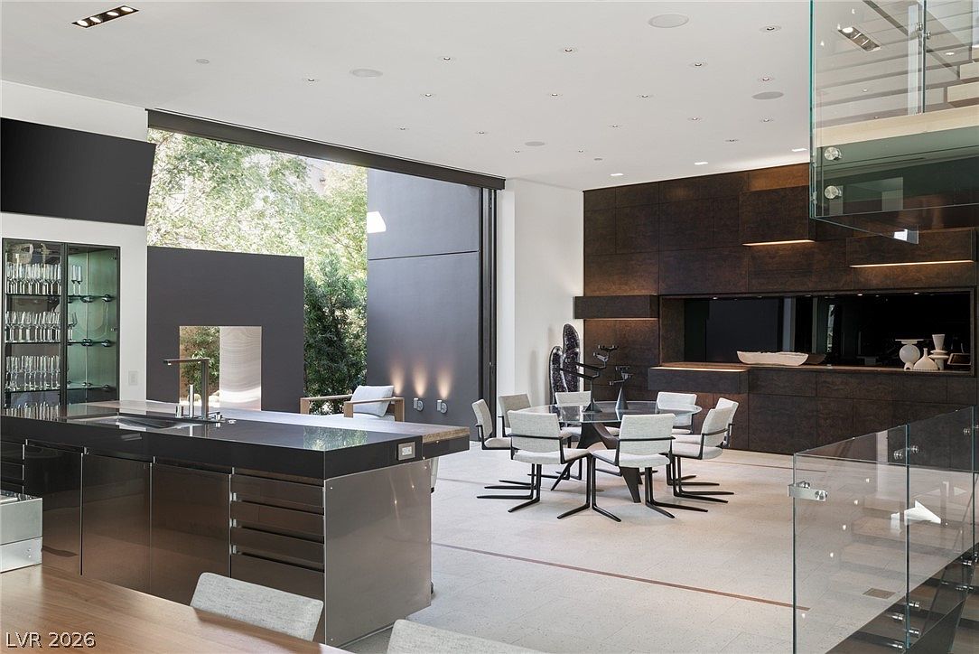 This expansive, modern open-concept space features a sleek kitchen island with a dark stone countertop and metallic cabinetry. A dining area with a glass-topped table and contemporary chairs sits adjacent to a large wall of glass that opens to an outdoor area. The room is characterized by high ceilings, recessed lighting, and a sophisticated, minimalist aesthetic with dark wood-paneled walls.