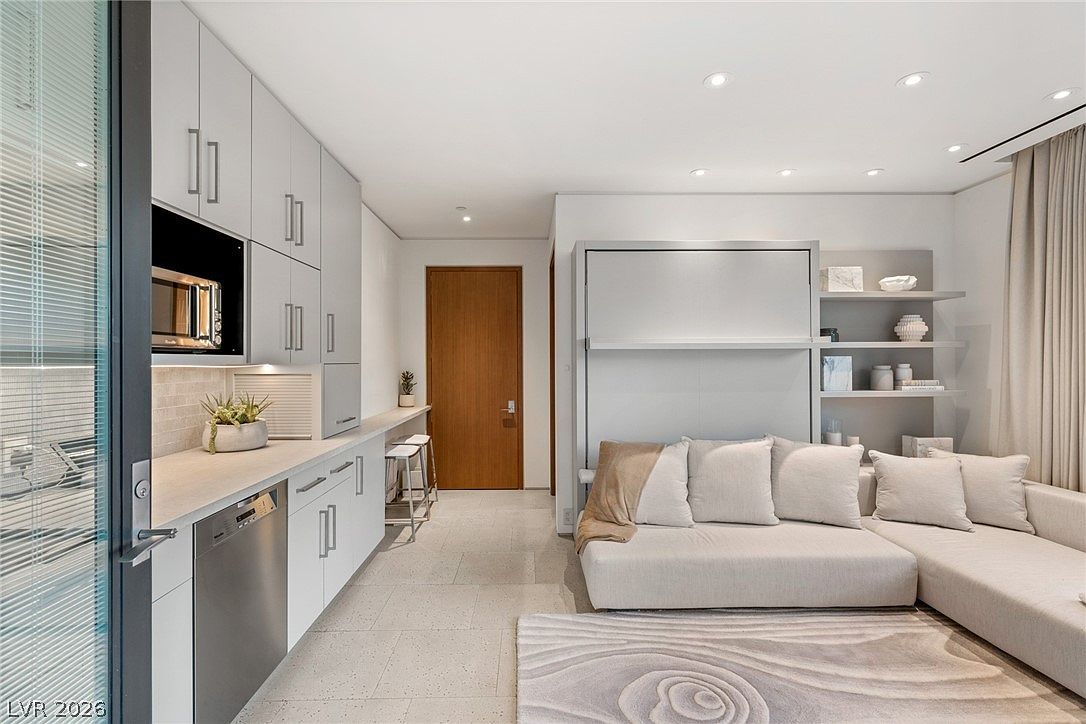 This modern studio-style space features a sleek, minimalist kitchen with light-toned cabinetry and integrated stainless steel appliances. A built-in counter serves as a breakfast bar, leading into a living area equipped with a wall-mounted bed unit and a contemporary sectional sofa. The room is finished with neutral-toned tile flooring and a textured area rug, creating a cohesive and functional living environment.