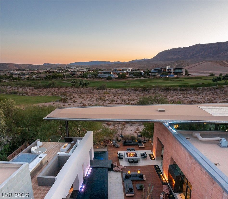 This high-angle aerial view captures a contemporary luxury residence featuring a sprawling outdoor living space with a sleek swimming pool and multiple lounge areas. The architecture emphasizes clean lines and a modern aesthetic, with a large cantilevered roof providing shade over the deck. Beyond the property, the landscape opens up to a golf course and desert terrain, framed by distant mountains under a soft sunset sky.