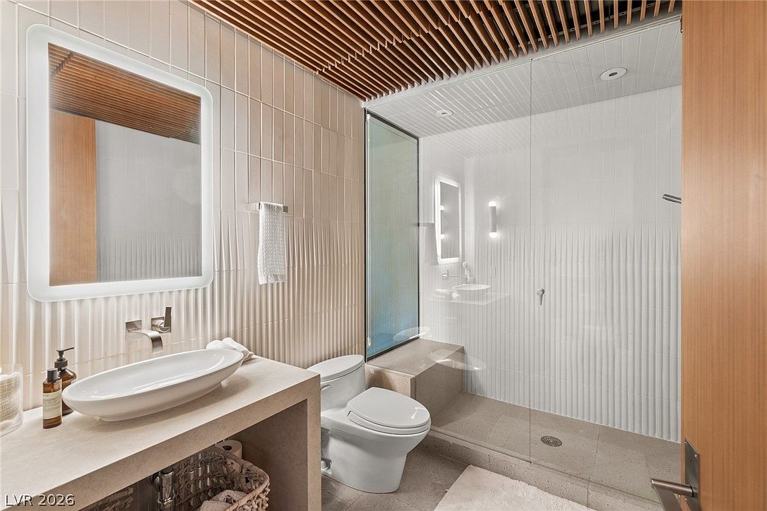 This modern bathroom features a sleek, wall-mounted vessel sink atop a custom stone vanity with an integrated lower shelf for storage. The space is defined by textured, vertical-ribbed wall tiles and a striking slatted wood ceiling that adds warmth and architectural interest. A glass-enclosed walk-in shower with a built-in stone bench completes the minimalist, spa-like aesthetic.
