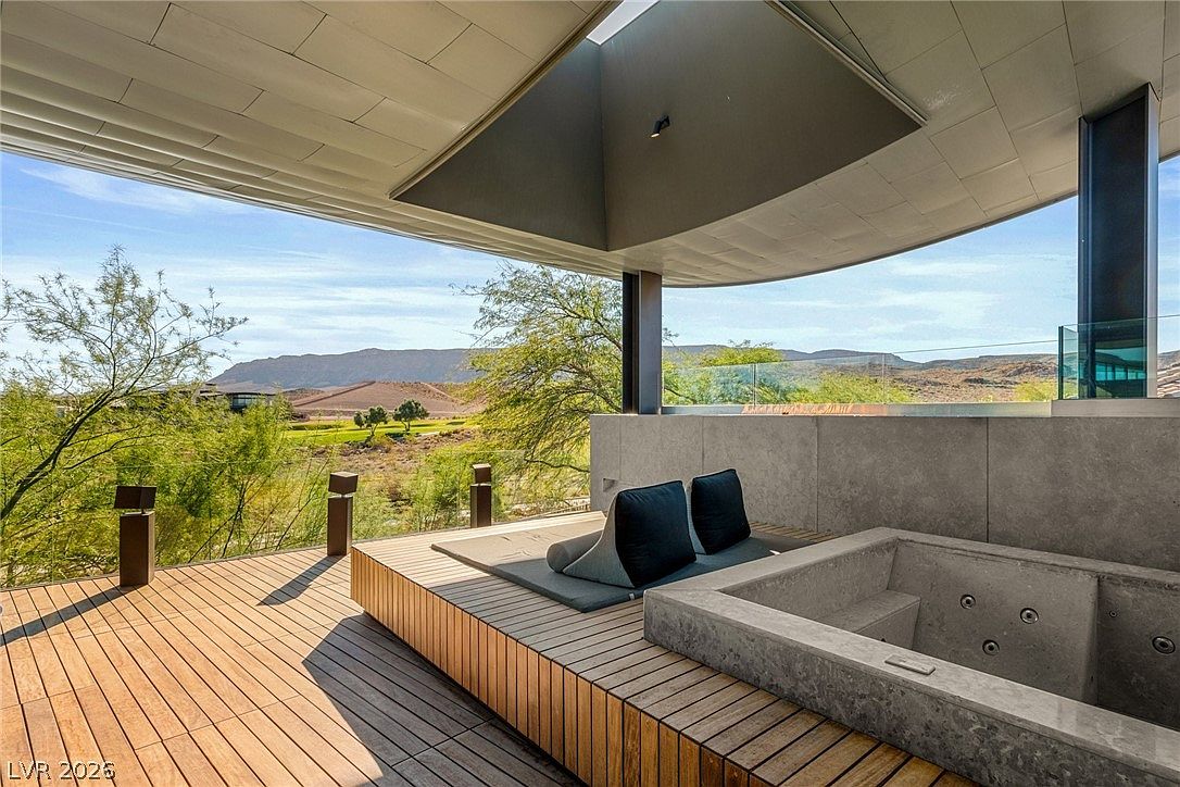 This modern outdoor terrace features a sleek, built-in concrete spa and a raised wooden deck platform with comfortable lounge seating. The space is sheltered by a dramatic, angular architectural overhang, framing expansive views of the desert landscape and distant mountains. The design emphasizes a seamless transition between indoor luxury and the natural environment.