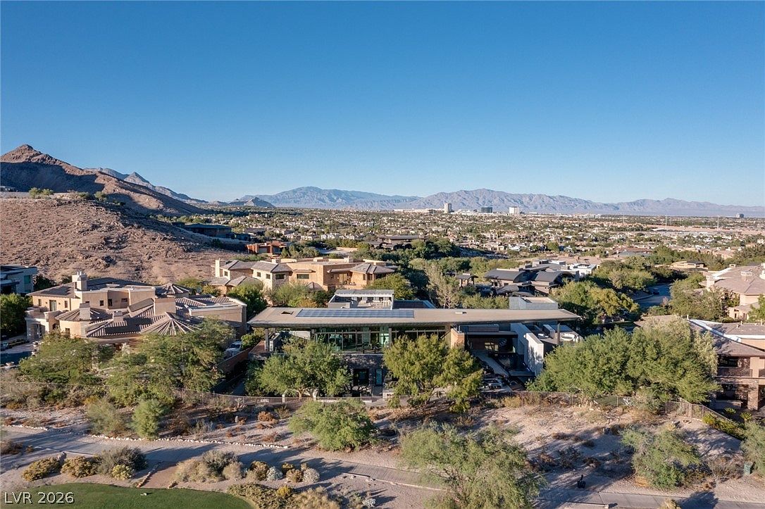 This high-angle aerial view captures a sprawling, modern luxury residence nestled in a desert landscape with dramatic mountain backdrops. The home features a flat roofline with integrated solar panels, expansive glass walls, and a multi-level design that blends seamlessly into the surrounding arid terrain. The property is situated within a quiet, upscale neighborhood characterized by desert landscaping and paved streets.