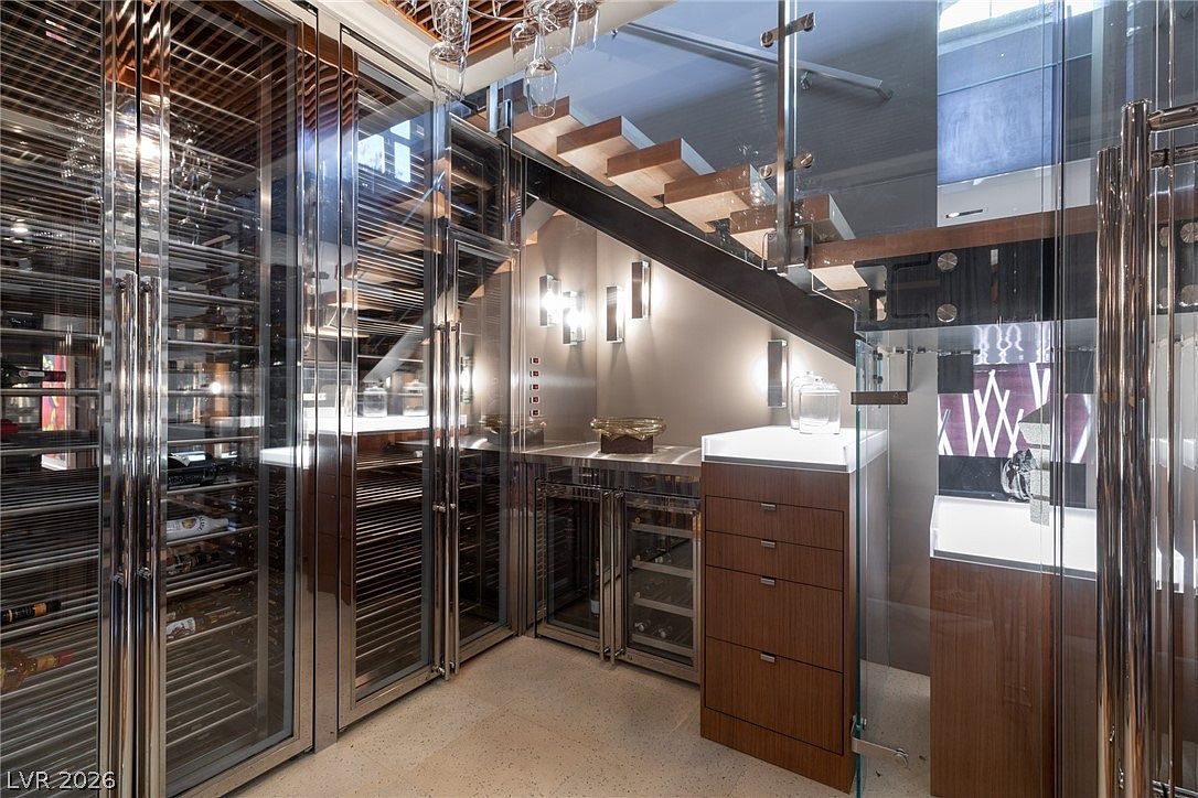 This sophisticated wine cellar features extensive glass-fronted climate-controlled storage units with polished metal frames. A floating staircase with glass railings ascends above a sleek service area equipped with under-counter beverage coolers and a wooden cabinet unit. The space is illuminated by modern wall sconces and recessed lighting, creating a luxurious and contemporary atmosphere.