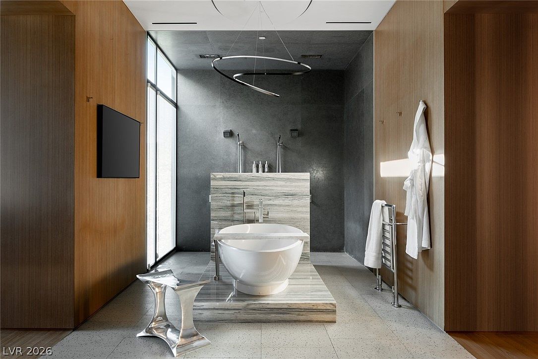 This luxurious primary bathroom features a central freestanding soaking tub set upon a raised stone platform, backed by a striking stone-clad partition wall. The space is framed by warm wood-paneled walls on either side, contrasting with the dark, textured stone wall behind the tub and the sleek, modern light fixture overhead. A wall-mounted television and a heated towel rack add functional elegance to this sophisticated, spa-like retreat.