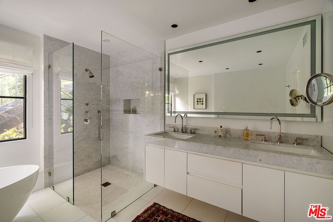 This is a bright and modern primary bathroom featuring a glass-enclosed shower with marble walls and a built-in niche. A large, illuminated mirror hangs above a double vanity with white cabinets and a marble countertop. The room is well-lit and exudes a clean, spa-like atmosphere.