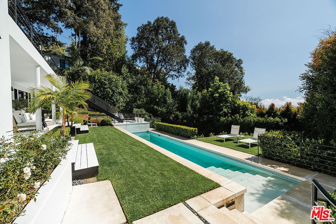 This image showcases a modern backyard featuring a long, narrow swimming pool with a light blue interior, surrounded by lush green grass and manicured hedges. A white bench and lounge chairs provide seating options, while a contemporary house with a black staircase is visible in the background. The scene exudes a sense of luxury and relaxation, perfect for outdoor entertaining.