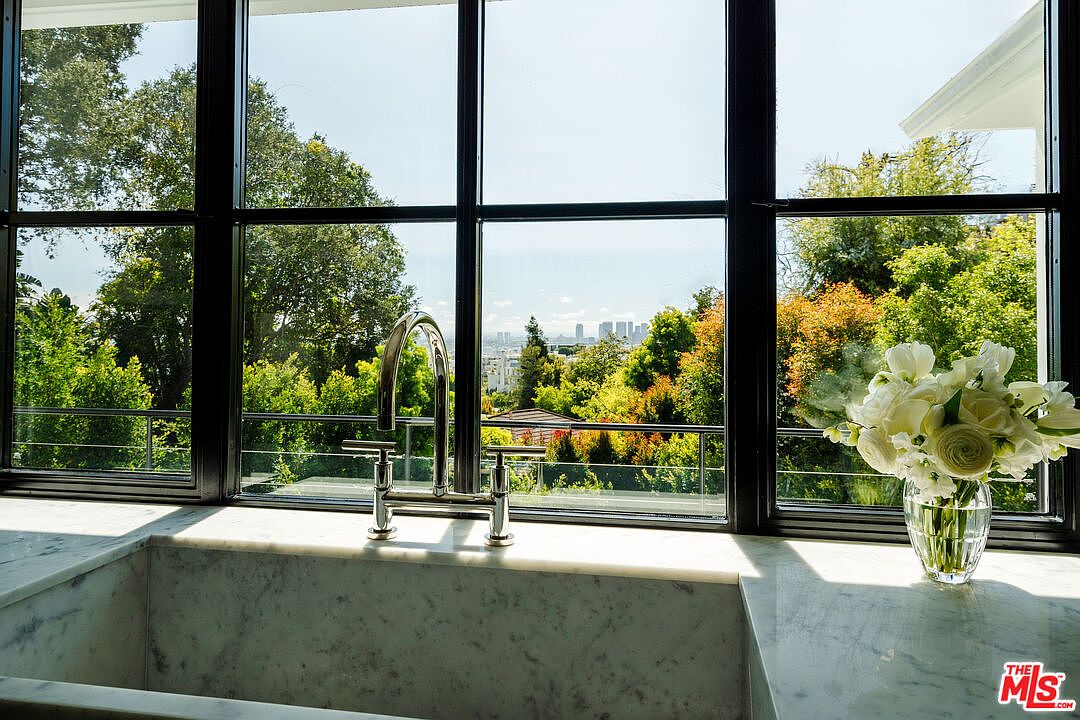 This image showcases a kitchen sink area with a large window offering a scenic view of lush greenery and distant city buildings. The sink is made of marble, complemented by a modern chrome faucet. A vase of white flowers adds a touch of elegance to the countertop, enhancing the overall aesthetic of the space.