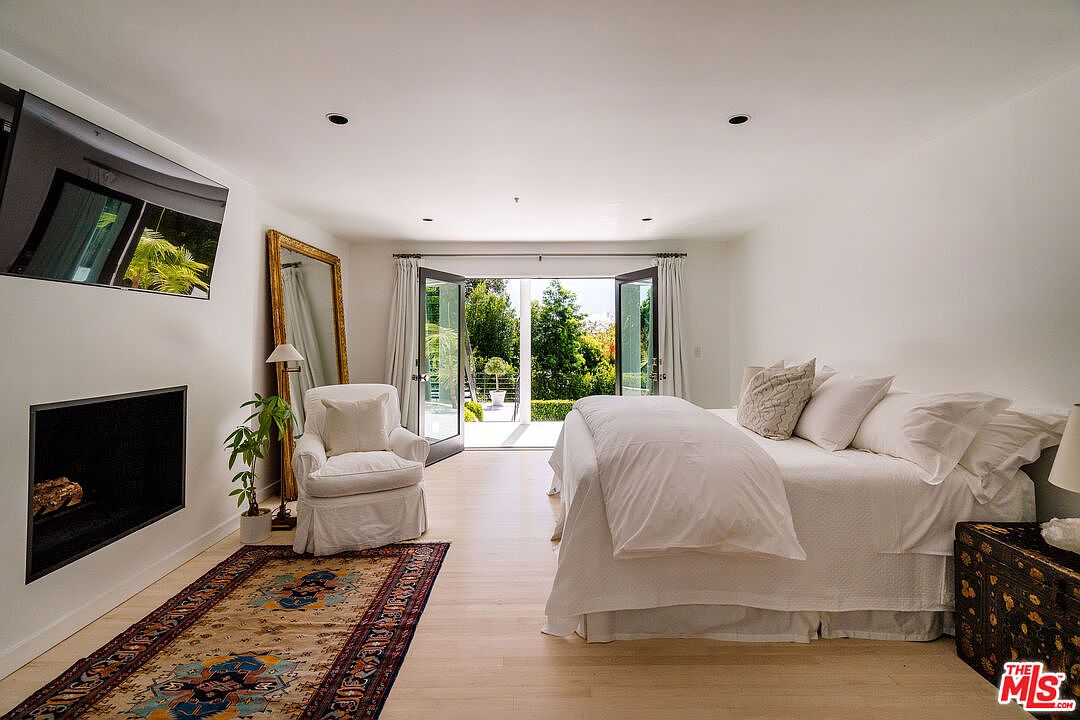 This is a bright and airy primary bedroom featuring a large bed with white linens, an accent chair, and a modern fireplace. French doors open to an outdoor area, bringing in natural light and views of the greenery. The room is decorated in a minimalist style with light wood floors and white walls, creating a serene and inviting atmosphere.
