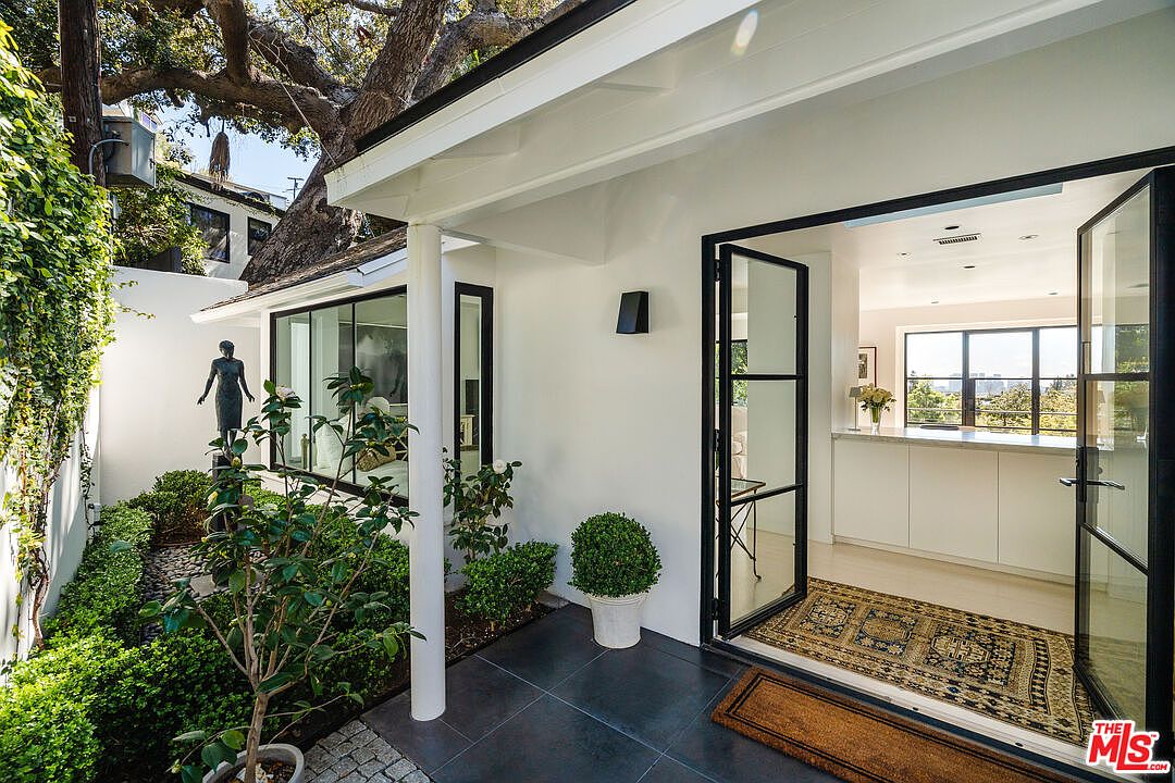 This is an inviting entryway to a modern home, featuring a well-manicured garden with a statue, black-framed windows, and a black-framed glass door leading into the interior. The exterior is painted in a clean white, contrasting with the black accents and the lush greenery. The perspective is from the outside looking in, showcasing the transition from the garden to the interior space.