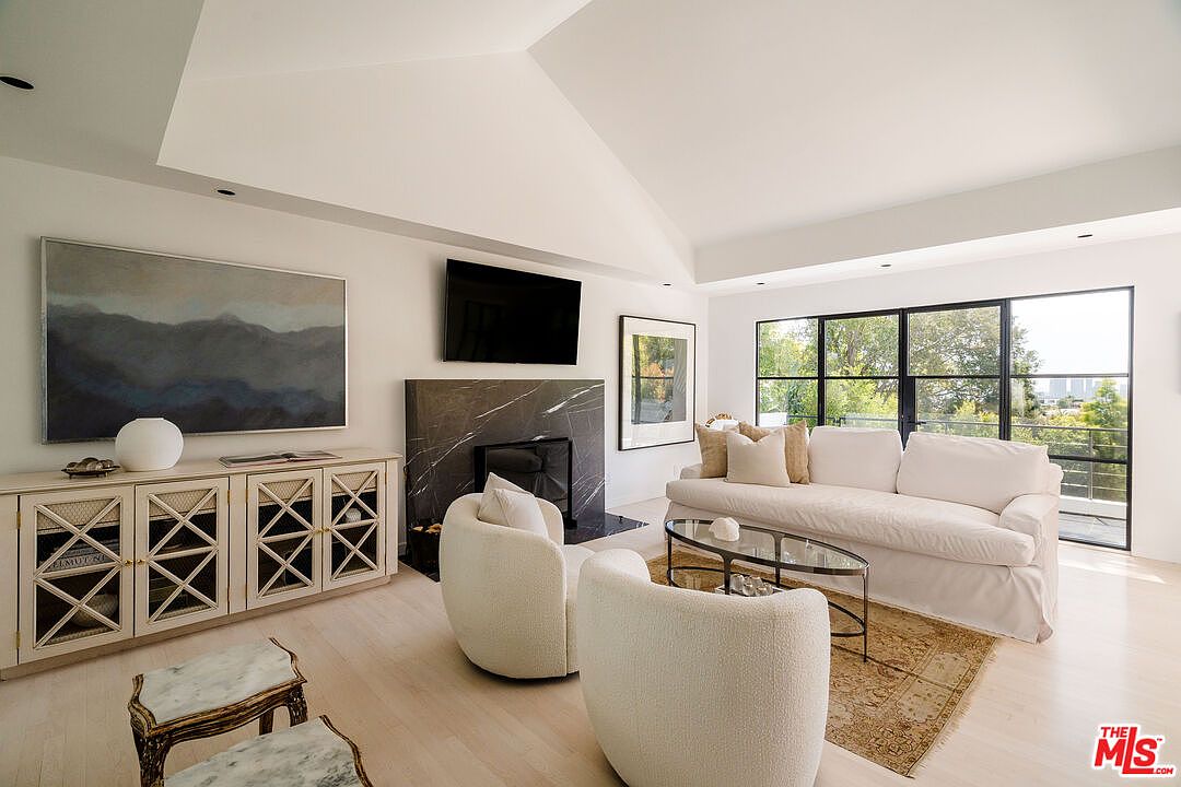 This is a bright and airy living room featuring a modern design with clean lines and a neutral color palette. The room includes a large painting, a flat-screen TV mounted above a marble fireplace, a white sofa, and two armchairs. Large windows offer a view of the outdoors, enhancing the sense of spaciousness and light.