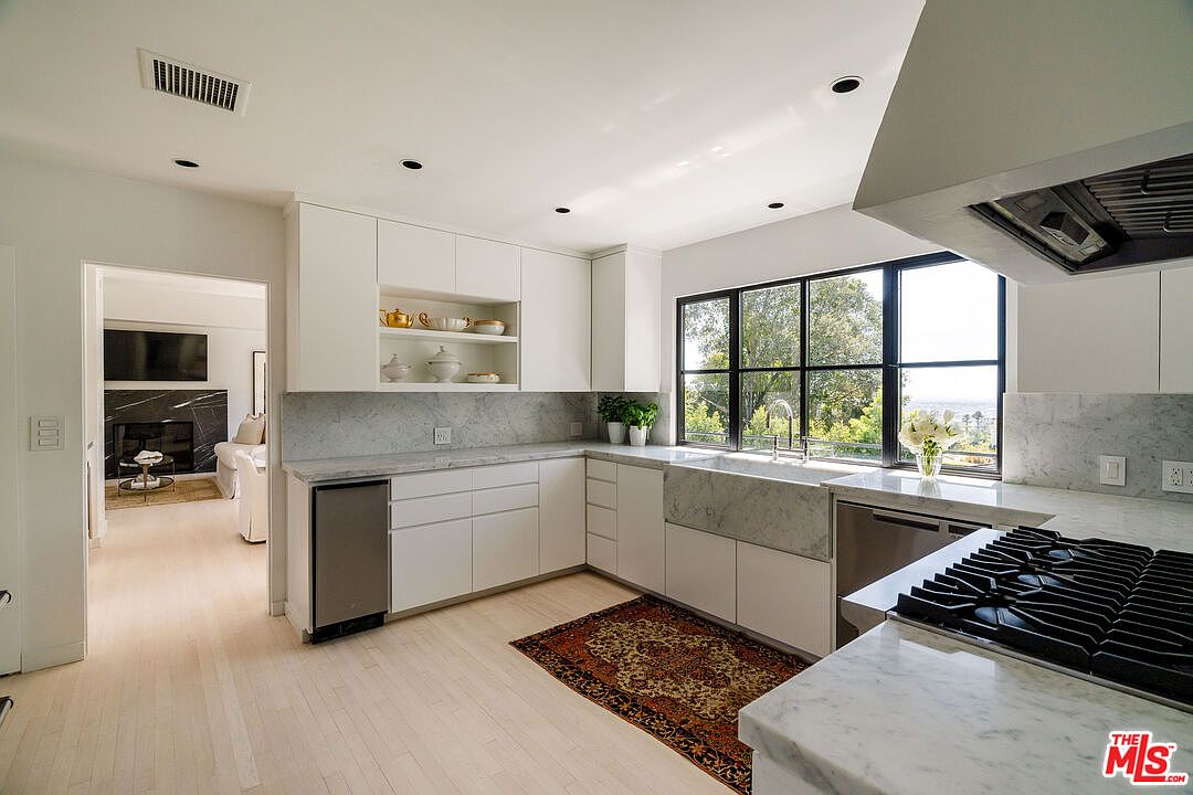 This is a bright and modern kitchen featuring white cabinetry, marble countertops and backsplash, and stainless steel appliances. A large window provides natural light and a view of the outdoors. The kitchen also includes a farmhouse sink and a patterned rug, adding a touch of warmth to the space.