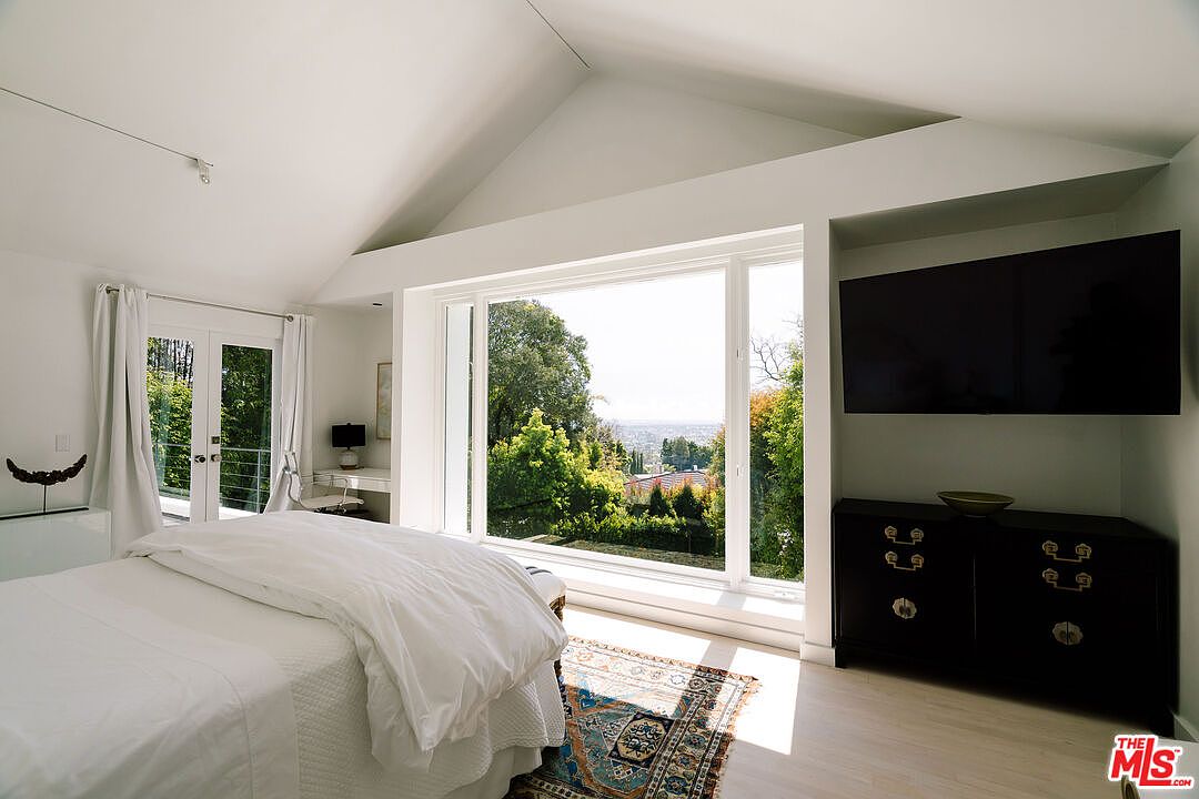 This is a bright and airy primary bedroom featuring a large window with a view of lush greenery and a city skyline. The room is decorated in a minimalist style with white walls, light wood floors, and a dark wood dresser with a large television mounted above it. A bed with white linens and a patterned rug add to the room's serene and inviting atmosphere.