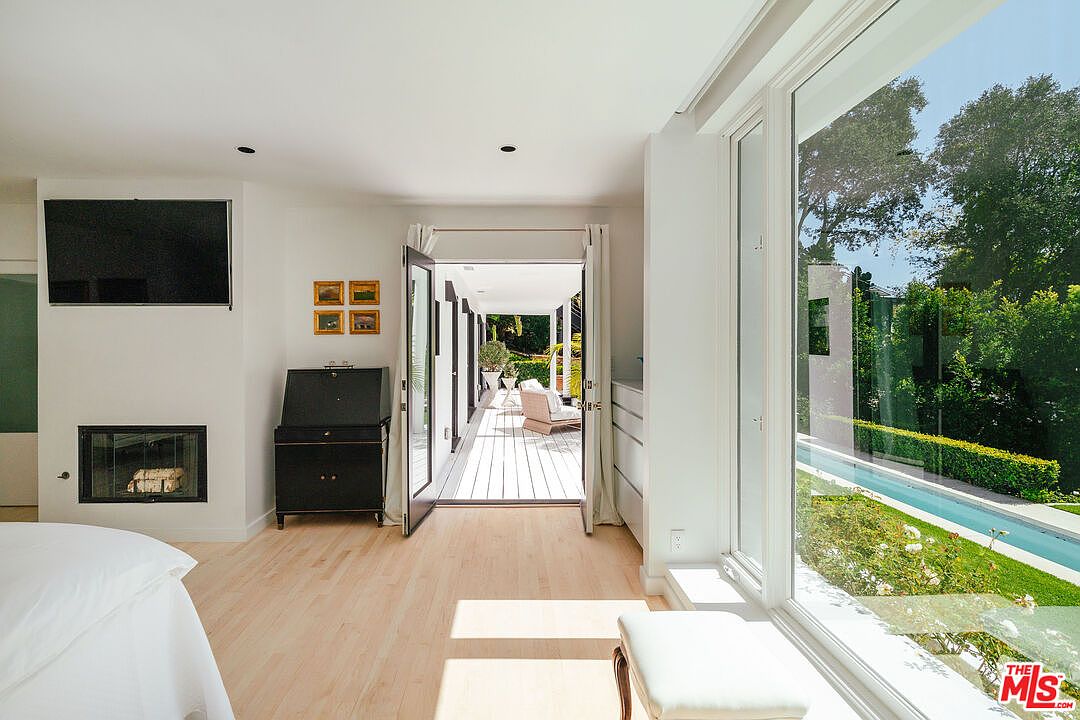 This is a bright and airy primary bedroom featuring a modern fireplace with a mounted television above, a dark wood secretary desk, and a large window overlooking a pool and lush greenery. The room has light hardwood floors and an open doorway leading to an outdoor deck, creating a seamless indoor-outdoor living experience. The overall aesthetic is clean, contemporary, and luxurious.