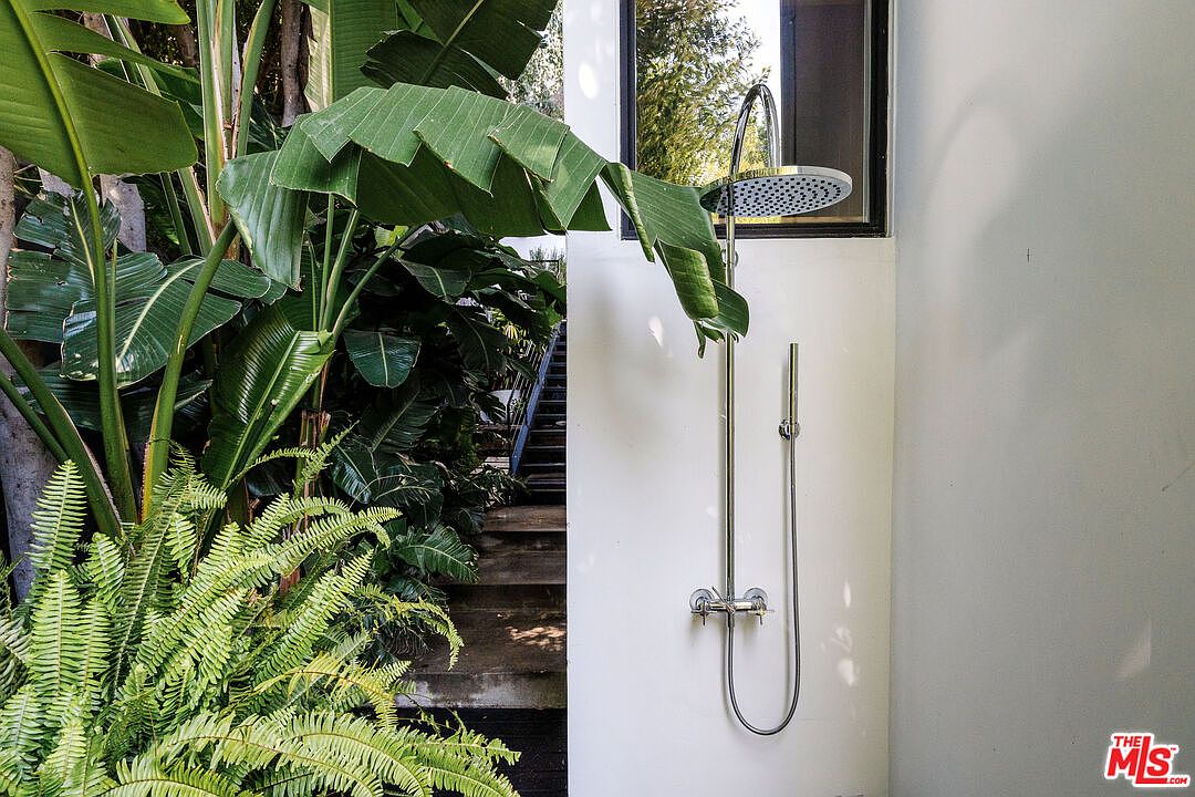 This image showcases an outdoor shower with a modern design, featuring a sleek, chrome fixture against a white wall. Lush greenery, including ferns and banana leaves, surrounds the shower area, creating a tropical and private ambiance. The presence of nearby stairs suggests easy access from an outdoor living space.