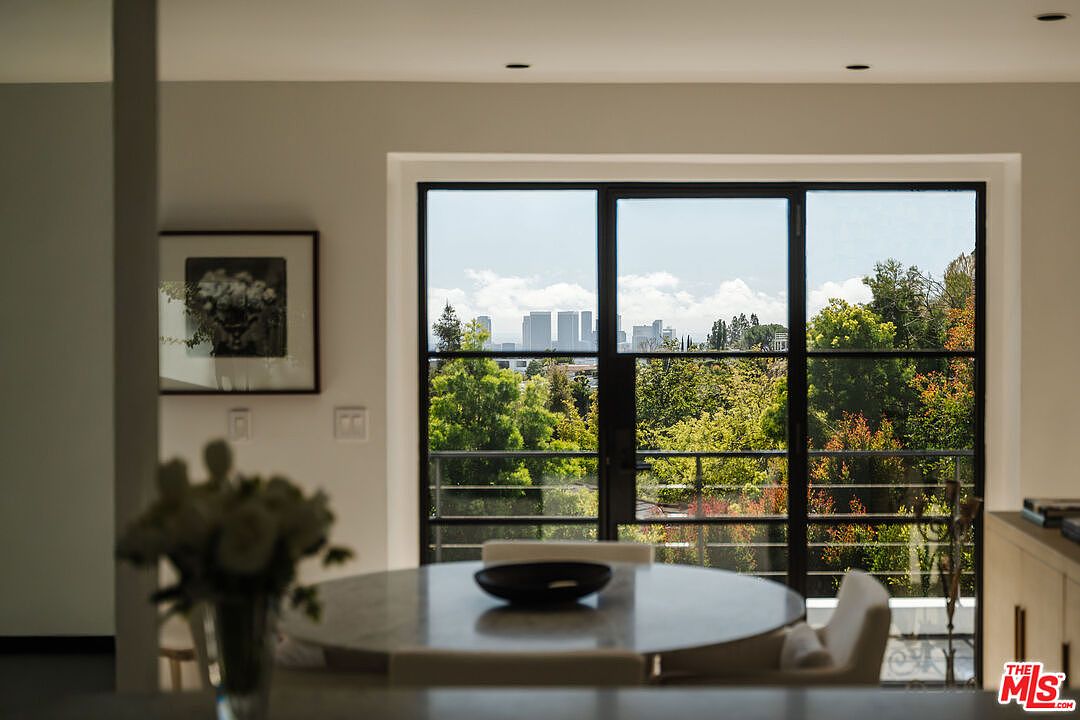 This interior shot showcases a dining area with a round marble table and modern chairs. A large window provides a view of lush greenery and distant city buildings, creating a serene backdrop. The room is bright and airy, with minimalist decor and a focus on natural light.