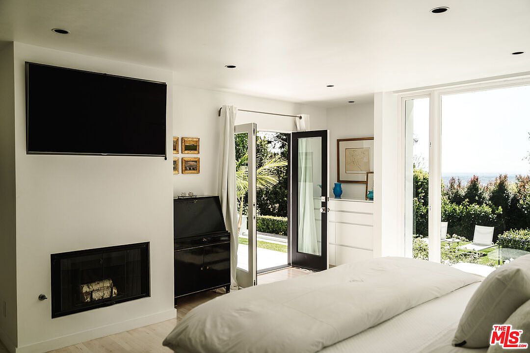 This is a bright and airy primary bedroom featuring a large bed with white linens, a modern fireplace with a mounted television above, and a black secretary desk. French doors open to a garden view, enhancing the room's connection to the outdoors. The room is decorated in a minimalist style with white walls and light wood floors, creating a serene and inviting atmosphere.