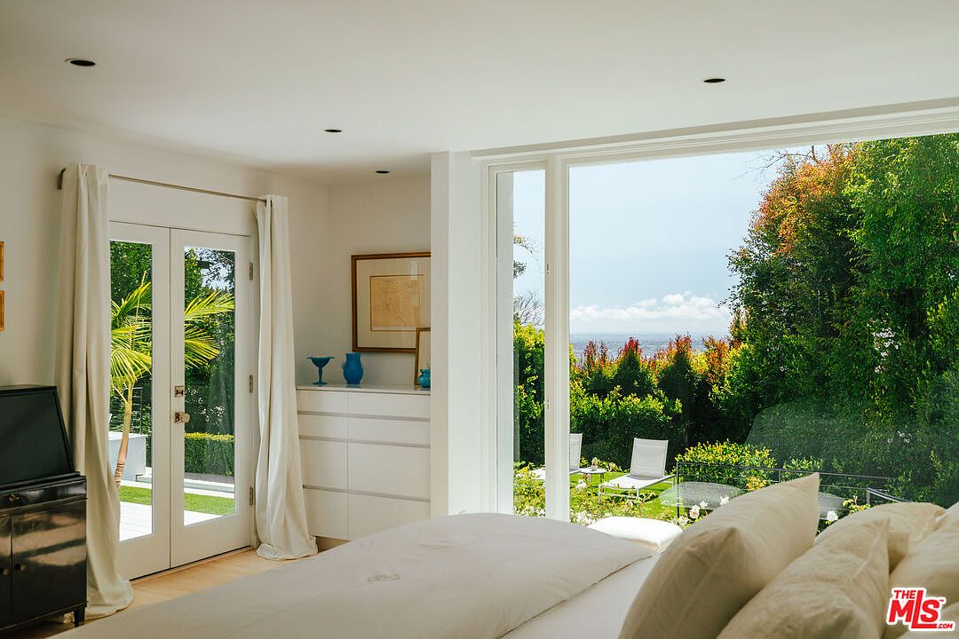 This primary bedroom features a bright and airy atmosphere with large windows offering views of a lush garden. The room includes a white dresser with decorative vases, and a set of French doors with white curtains. The bed is neatly made with white linens, creating a serene and inviting space.