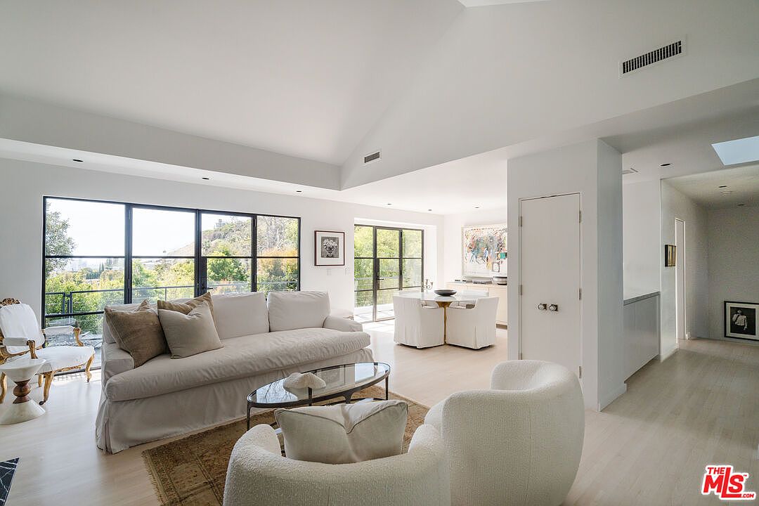 This is an interior shot of a bright and airy living room. The room features a large sofa, a comfortable armchair, and a glass-topped coffee table, all arranged on a neutral-toned rug. Large windows offer views of the surrounding greenery, and the overall aesthetic is modern and minimalist with white walls and light wood floors.