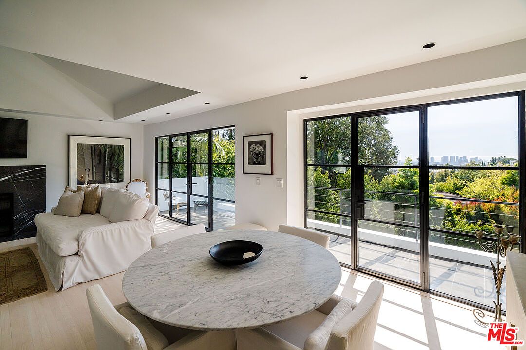 This interior shot showcases a bright and airy dining room with a modern aesthetic. A round marble table is the centerpiece, surrounded by upholstered chairs. Large black-framed windows offer expansive views of lush greenery and a distant cityscape, enhancing the room's connection to the outdoors.