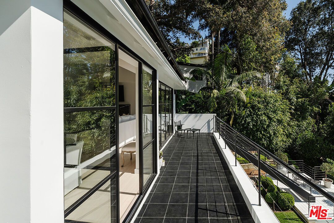 This image showcases a stylish outdoor balcony or patio area. The space features black tiled flooring, modern black-framed glass doors leading indoors, and a sleek black railing. The area is surrounded by lush greenery, creating a private and serene atmosphere, perfect for relaxation or entertaining.